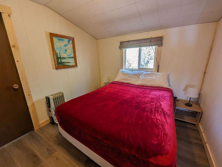A welcoming master bedroom featuring a queen-sized bed with a deep red blanket under a large window. A landscape painting adds character to the wall.