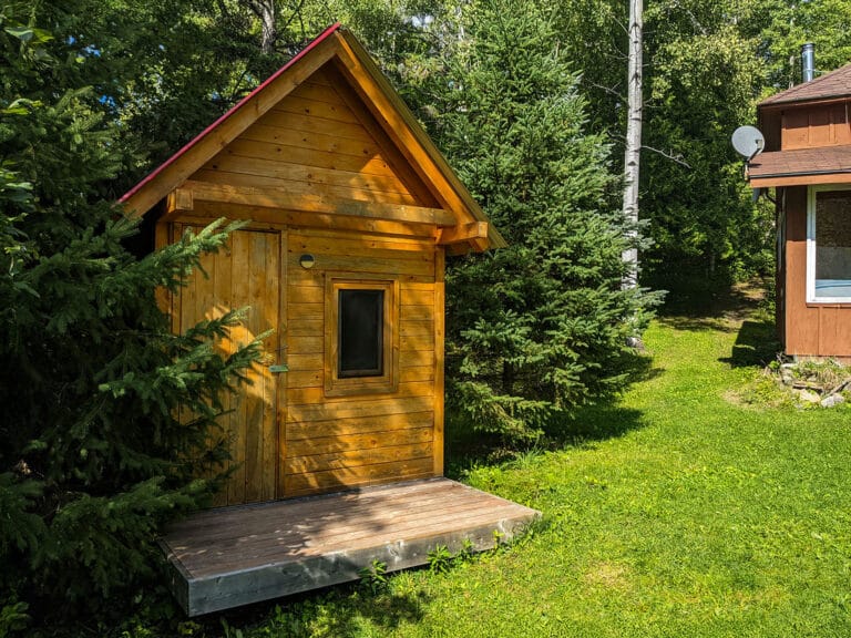 A small, red-roofed wooden bunkie tucked among the trees. Its natural wood exterior and setting make it an ideal, private getaway spot.