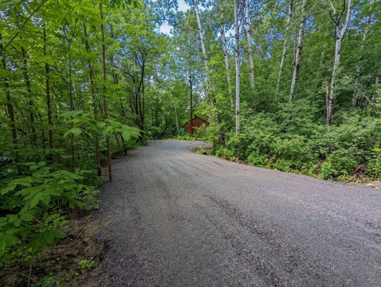 A winding gravel driveway leads through a forested area with lush green trees on either side, providing a natural entrance to the cottage.