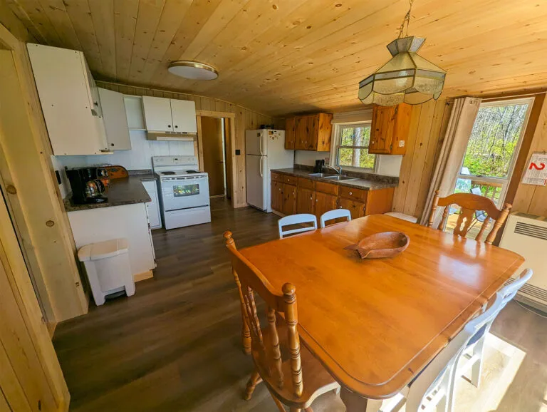 A full kitchen setup with wooden cabinets and countertops. The dining area has a large window showcasing a serene lake view. The ceiling is wood-paneled, creating a warm and rustic feel.