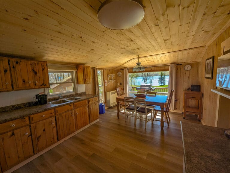 A spacious kitchen with a wood-paneled ceiling, white and wooden cabinets, and a dining table seating six. Natural light fills the room from large windows with a view of the greenery outside.